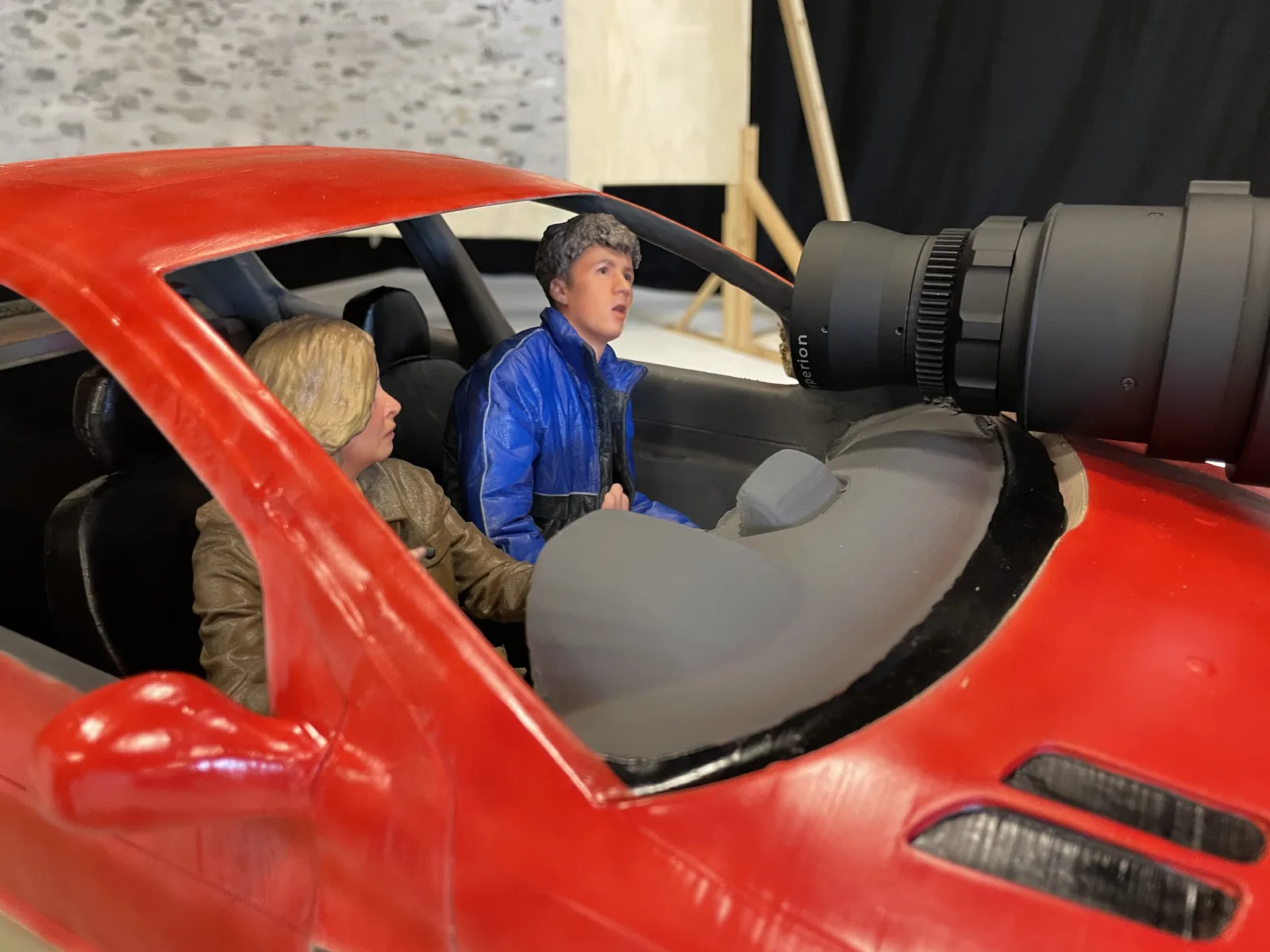 Painted miniature figures seated inside the red model car beside a camera lens